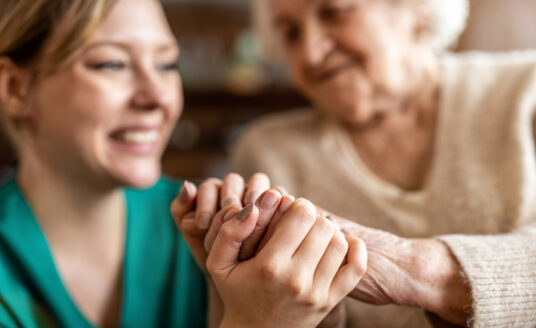 A young woman and older woman hold hands and smile in a hospice care facility, despite hospice care myths they may have heard.