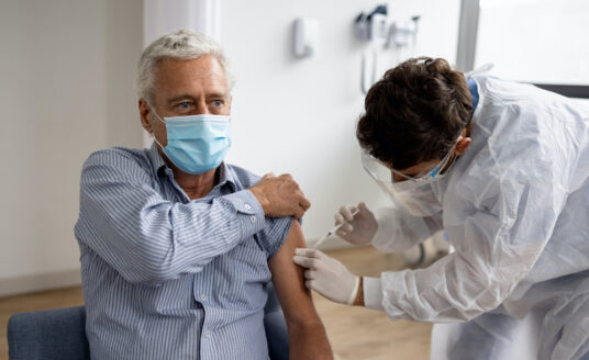 A senior man receives the Covid 19 booster with his sleeve rolled up and a mask on in a doctor's office.