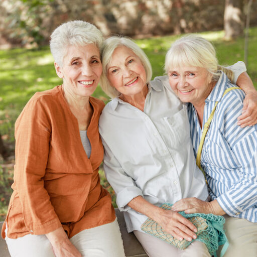 Three senior women embracing each other on a bench outside, maintaining their friendship for a healthy heart.