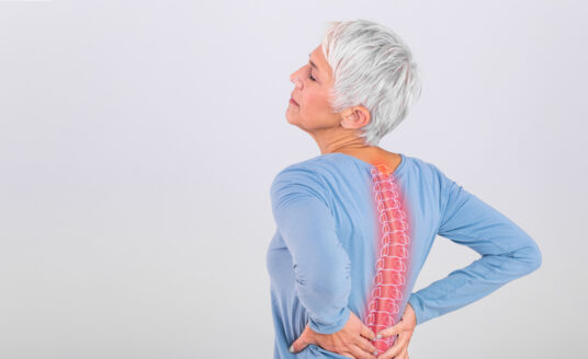 A senior woman in a blue shirt holds her back in pain, with a visual representation of her spine showing changes to posture.