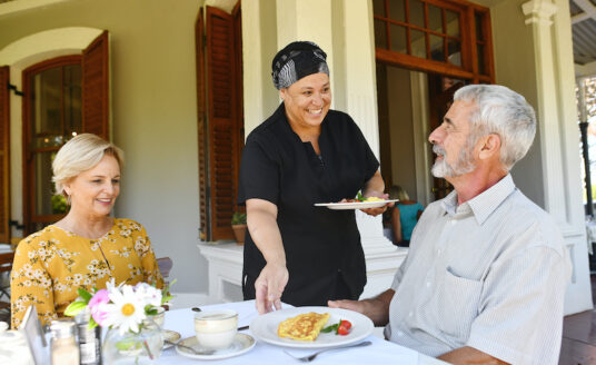 An older woman and man sit at a table and are served food while dining at an independent living community.