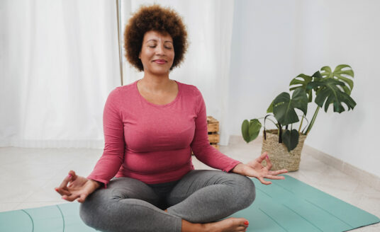 An older woman reaps the benefits of yoga as she sits on a yoga mat in a meditation pose.