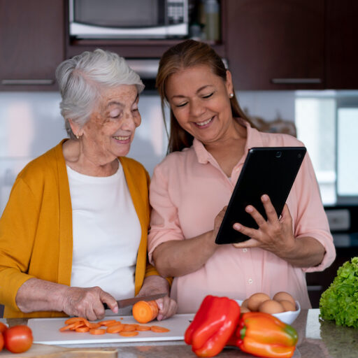A middle-aged female caregiver and a senior-aged woman reap the benefits of cooking by creating a meal together in a kitchen.