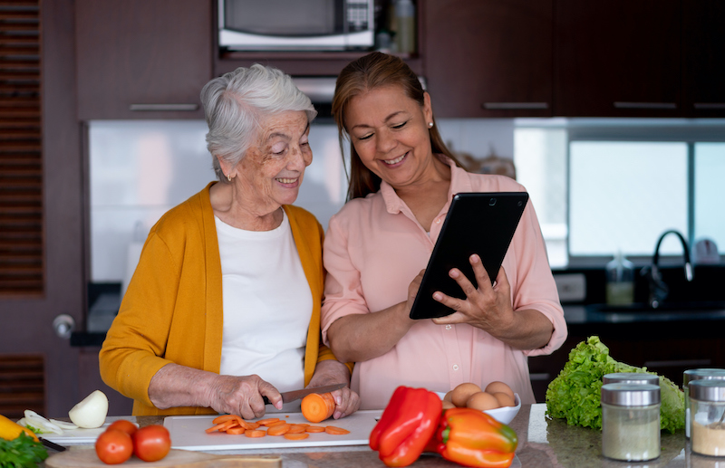 A middle-aged female caregiver and a senior-aged woman reap the benefits of cooking by creating a meal together in a kitchen.