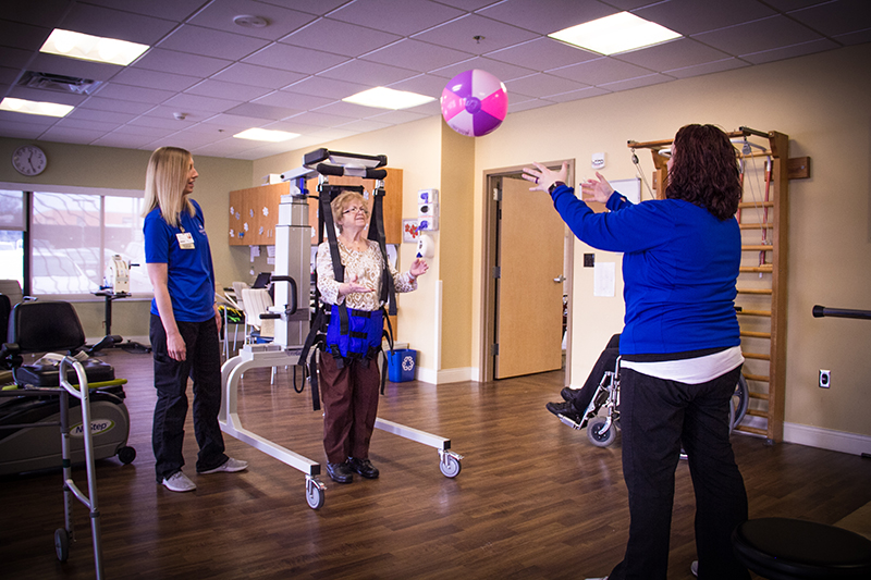 A senior woman in a standing frame throws a weighted exercise ball to a female physical therapist