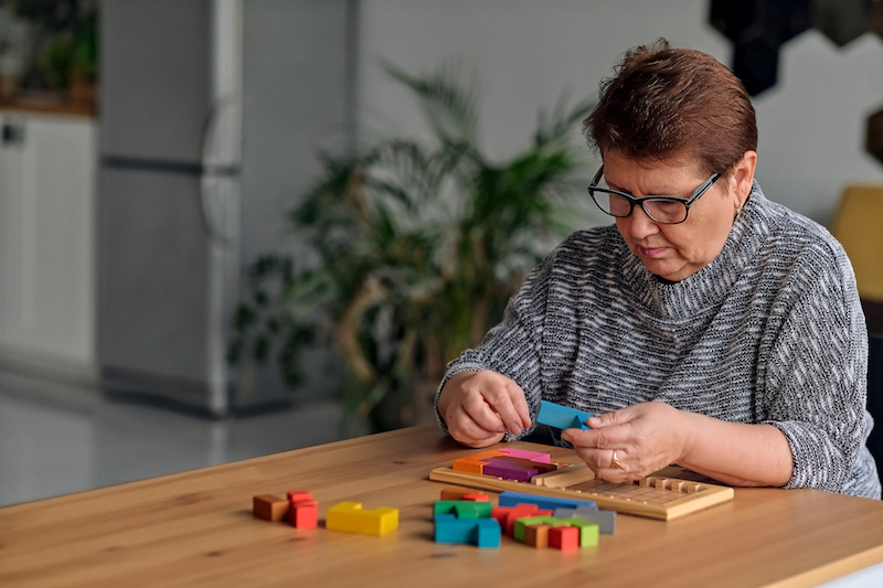 An older woman puts together a colorful wooden puzzle on a dining room table as one of many brain exercises.