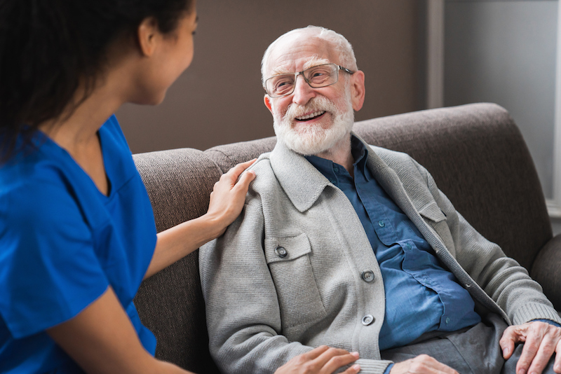 A young female nurse in a memory care community attends to a senior gentleman sitting on a couch.