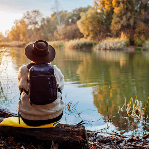 A woman wearing a sweater and a hat sits at a riverbank, realizing the importance of nature.