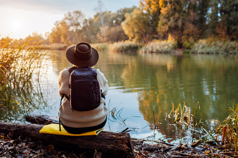 A woman wearing a sweater and a hat sits at a riverbank, realizing the importance of nature.
