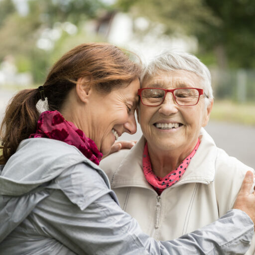 A senior woman and her younger female caregiver embrace during a walk outside, smiling as they embrace humor for family caregivers.