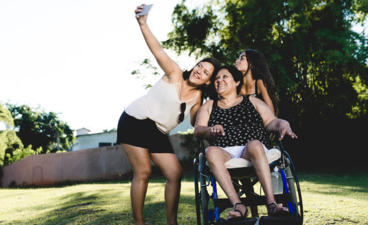 older women in a wheelchair taking a picture with two younger women in the park while doing activities for seniors