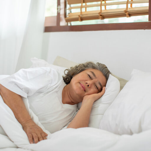 A senior aged woman lays in bed underneath the covers and sleeps, knowing that sleep keeps your mind sharp.