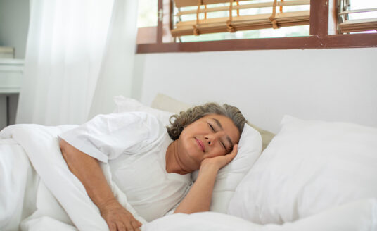 A senior aged woman lays in bed underneath the covers and sleeps, knowing that sleep keeps your mind sharp.