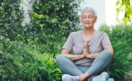 A senior woman reduces work stress by practicing meditation outside on a yoga mat.