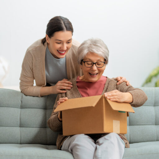 A senior woman happily opening a care package while her adult daughter leans over her shoulder.