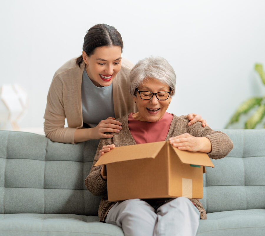 A senior woman happily opening a care package while her adult daughter leans over her shoulder.