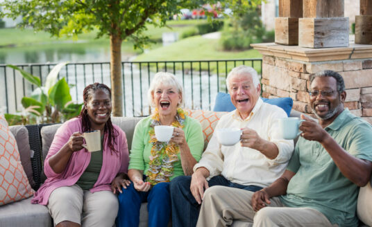 A group of happy elder men and women sitting on a couch in the outdoors having a drink in a cup