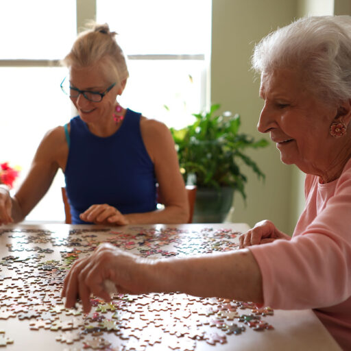 A smiling older woman working on a puzzle with her middle-aged daughter.