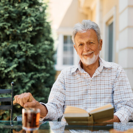 A smiling older man reads a book on a patio in an independent living community.