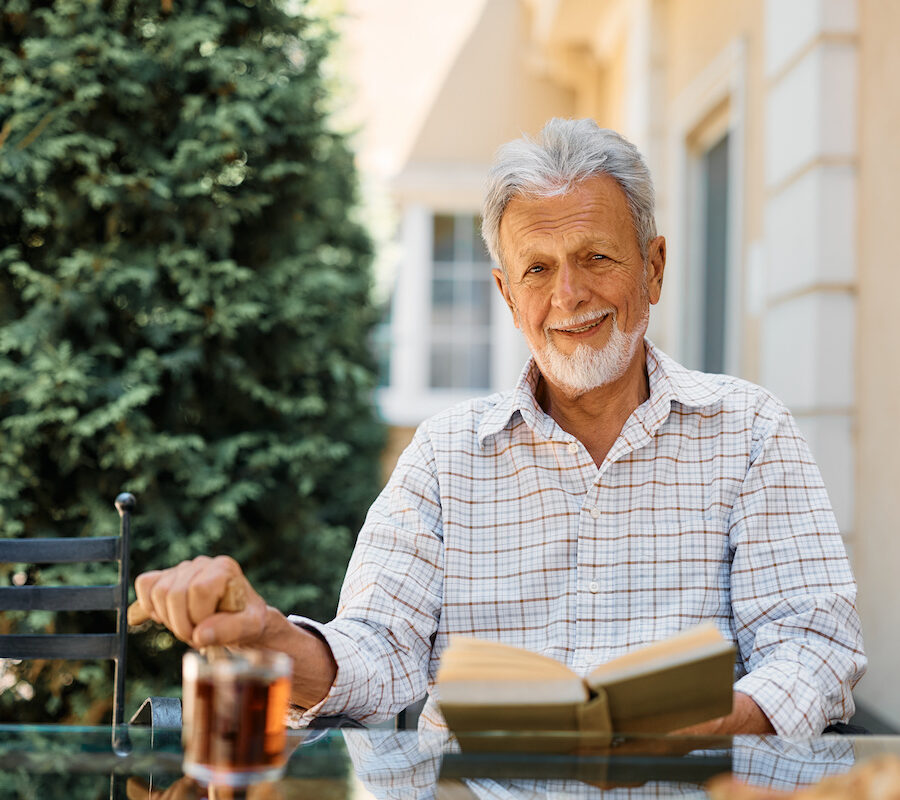 A smiling older man reads a book on a patio in an independent living community.