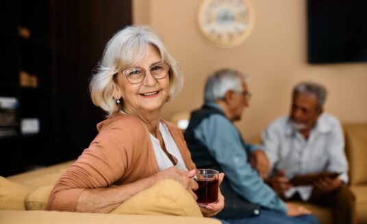 Smiling senior woman enjoys in cup of tea at senior living house and looks at camera