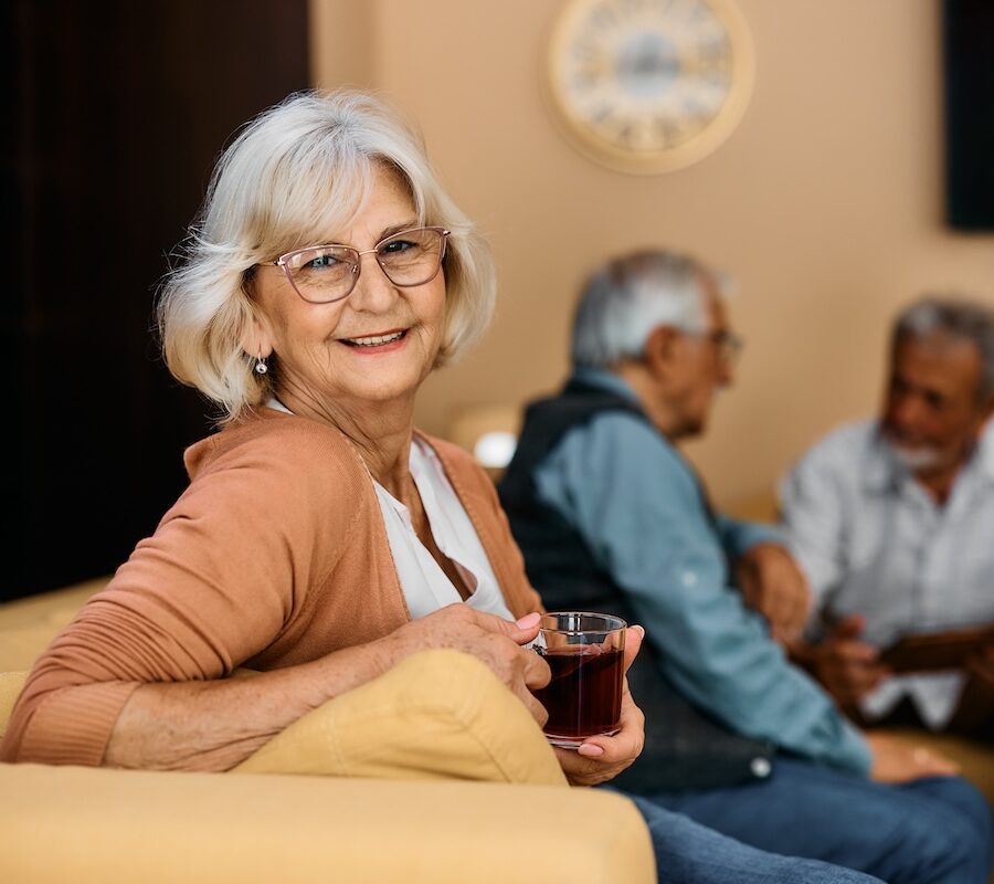 Smiling senior woman enjoys in cup of tea at senior living house and looks at camera