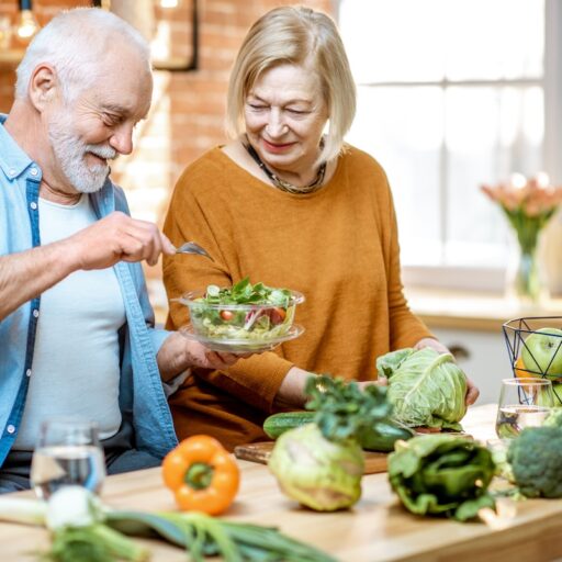 Cheerful senior couple eating salad standing together with healthy food on the kitchen at home.