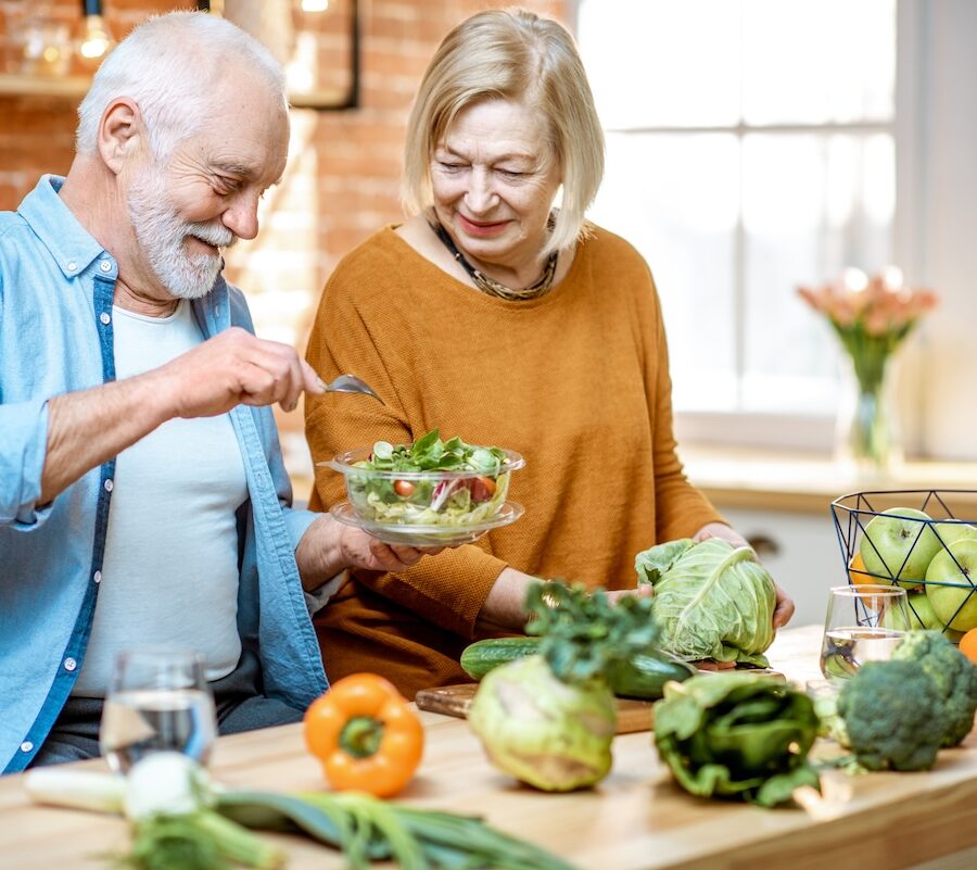 Cheerful senior couple eating salad standing together with healthy food on the kitchen at home.