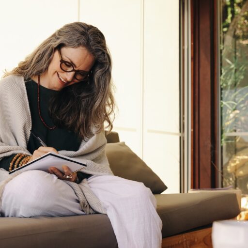 A woman sitting on a sofa, writing in a notebook, with a window view of rocks and greenery in the background.