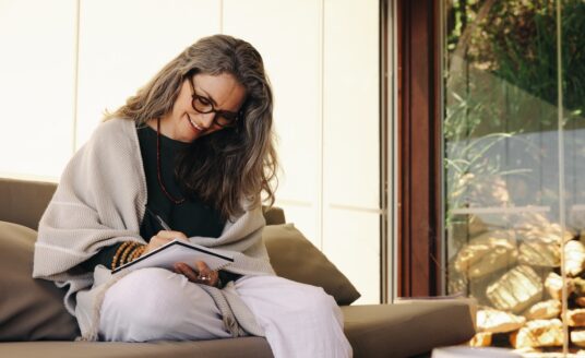 A woman sitting on a sofa, writing in a notebook, with a window view of rocks and greenery in the background.