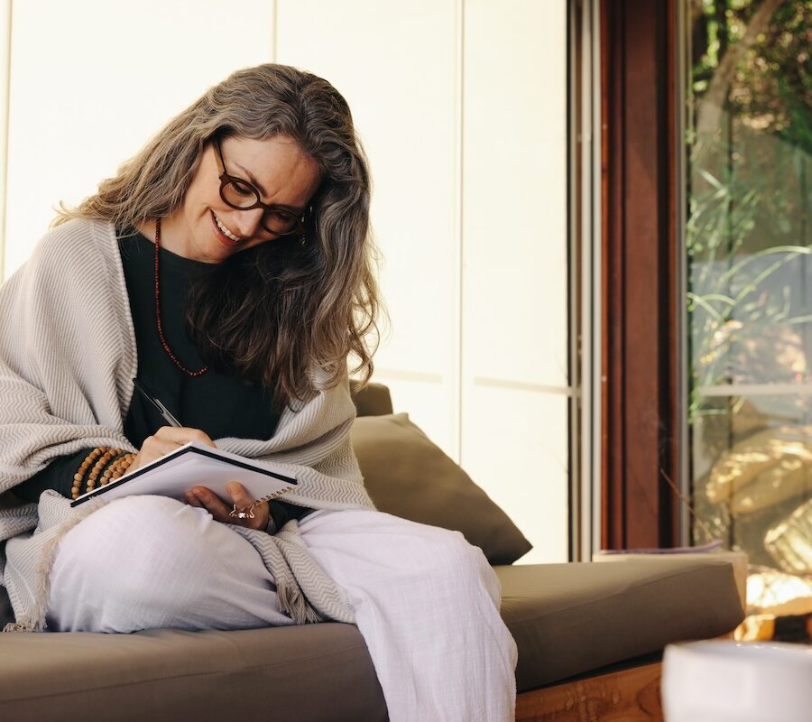 A woman sitting on a sofa, writing in a notebook, with a window view of rocks and greenery in the background.