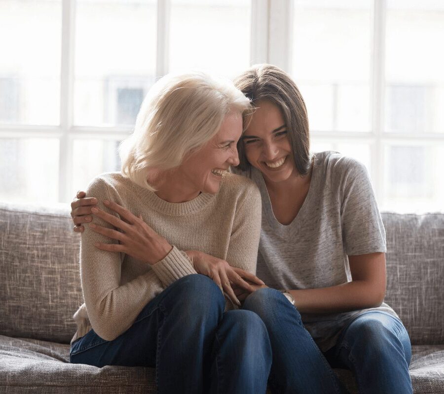 Two women sitting on a sofa, laughing together in front of large windows.
