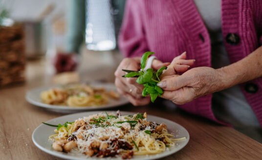 Senior woman snipping basil leaves from herb garden onto pasta