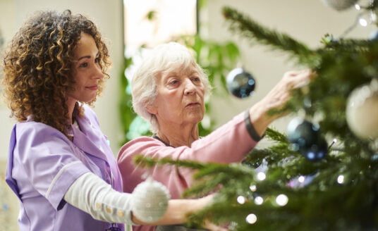 Home health care worker helping senior woman decorate Christmas tree for the holidays