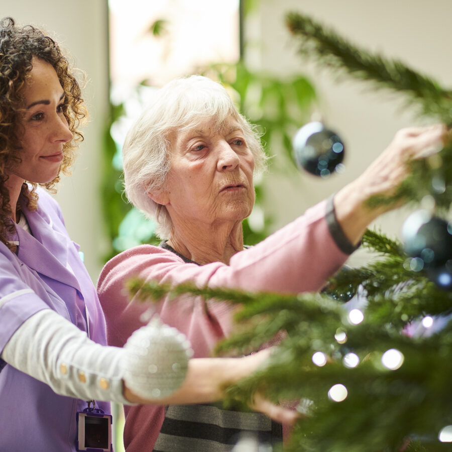 Home health care worker helping senior woman decorate Christmas tree for the holidays