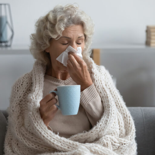 Senior woman blowing nose into tissue while wrapped in blanket, using flu season tips to feel healthy again
