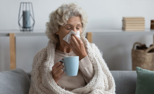 Senior woman blowing nose into tissue while wrapped in blanket, using flu season tips to feel healthy again