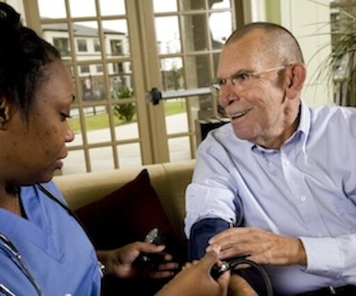 Nurse in blue scrubs checks an elderly man's blood pressure at a home setting.