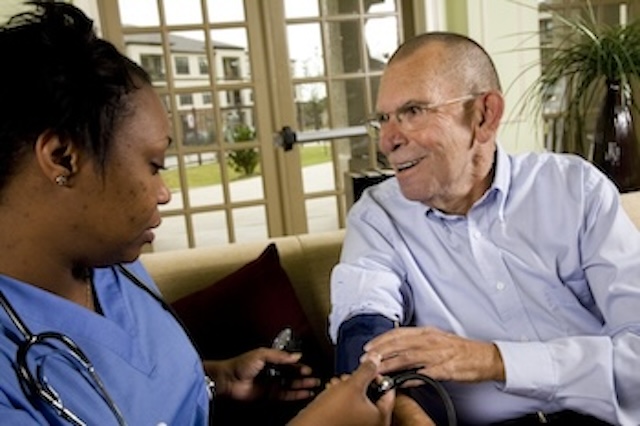 Nurse in blue scrubs checks an elderly man's blood pressure at a home setting.