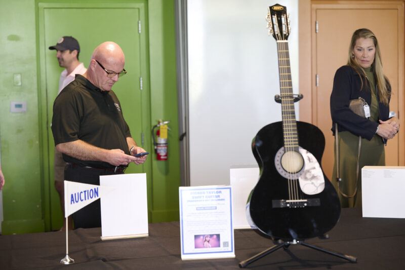 Black acoustic guitar on a stand at an auction table, with an AUCTION sign; a man in a black polo looks at his phone nearby, a woman in green stands in the background.