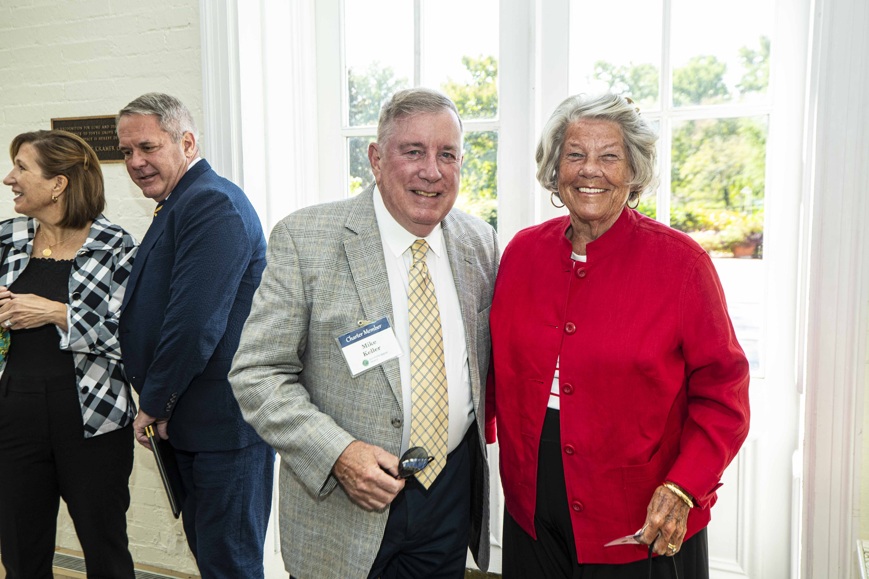 Smiling older man in a light plaid blazer and a red-jacketed woman stand together near a bright window, posing for a photo with others in the background.