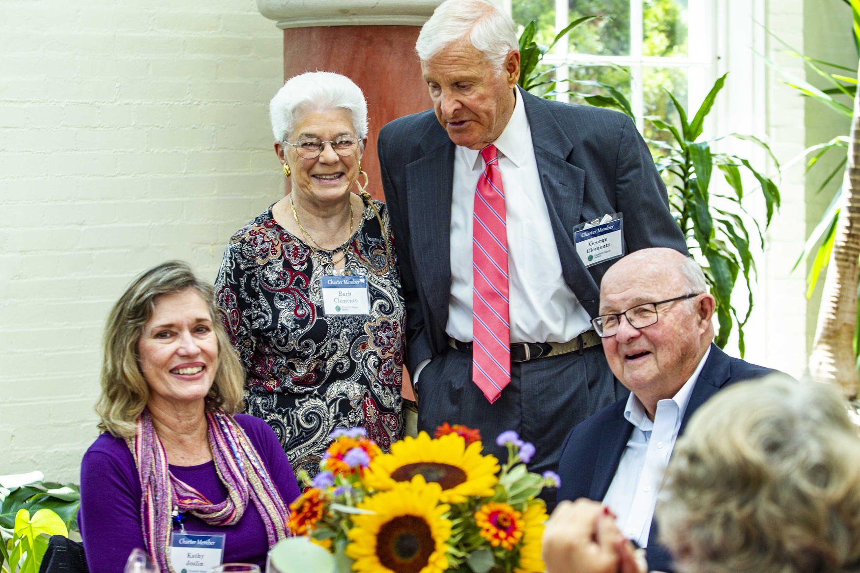 Group of smiling older adults at a social event, wearing name badges, gathered around a table with bright sunflowers in the foreground.