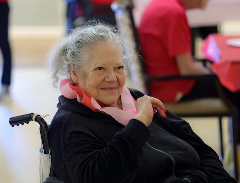 Older woman in a wheelchair, gray hair, wearing a black jacket and pink scarf, smiling softly in an indoor setting.