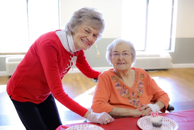 Caregiver in a red sweater helping an elderly woman at a table with cupcakes.