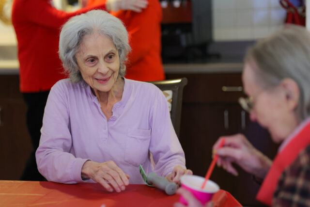 Older woman in a lavender shirt smiling while folding paper at a red table in a communal setting, with others in the background.