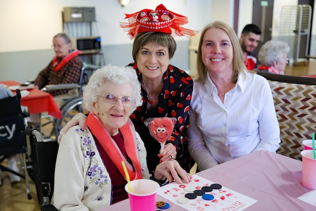 Elderly woman in a wheelchair at a table with two smiling companions; one wears a red dragon hat and holds a heart decoration in a festive room.