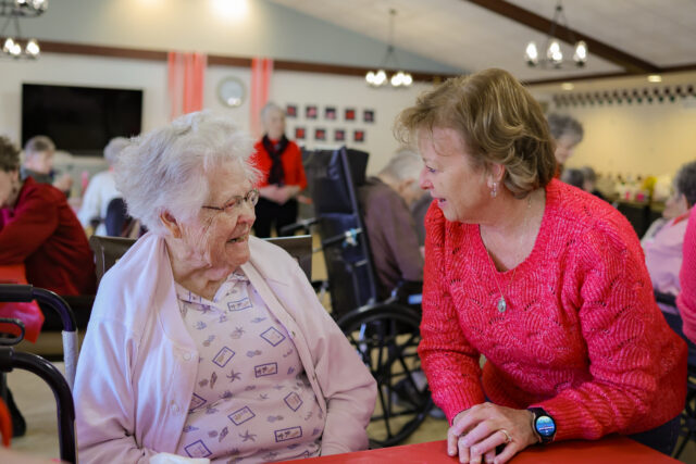 Two elderly women chatting at a table in a bright communal room; one in a pink sweater leans toward the other in a light jacket.