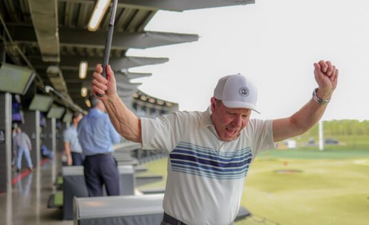 Man in a white cap and striped polo celebrates with arms raised at a golf driving range, holding a club