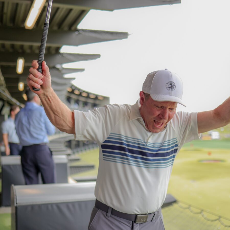 Man in a white cap and striped polo celebrates with arms raised at a golf driving range, holding a club