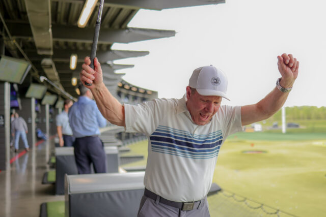 Older man at a golf driving range raises his club in celebration, wearing a white cap and striped polo shirt.